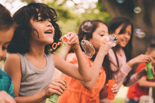 Little Boy Having Fun With Friends In Park Blowing Bubbles