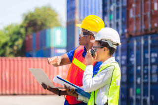 photo - Workers in Hardhats and Safety Vests Holding Laptop Checklist and Radio with Containers in Background