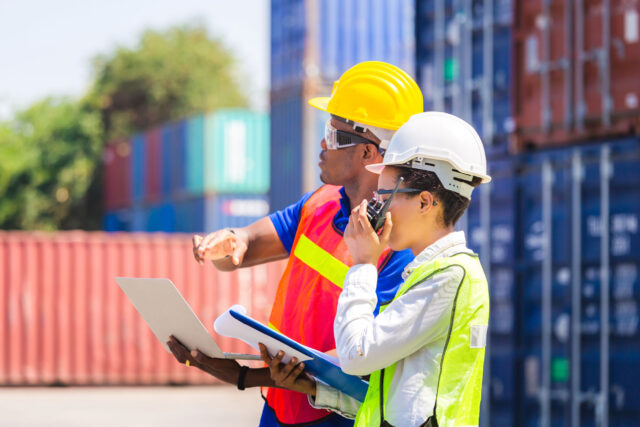 photo - Workers in Hardhats and Safety Vests Holding Laptop Checklist and Radio with Containers in Background