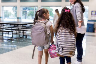 photo - Teacher Guides Elementary Age Girls Wearing Backpacks into Cafeteria