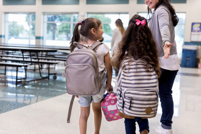 photo - Teacher Guides Elementary Age Girls Wearing Backpacks into Cafeteria