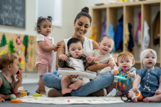 photo - Childcare Provider Sits with a Large Group of Babies as She Read Them a Book