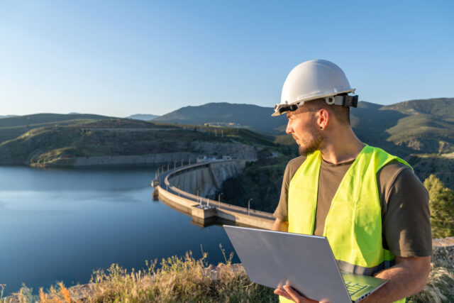 photo - Engineer Inspecting a Reservoir Dam and Holding Laptop