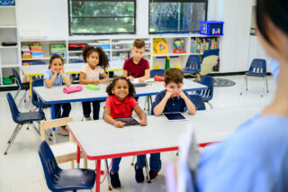 photo - Elementary School Students Sitting at Tables in Classroom and Looking Up at the Teacher