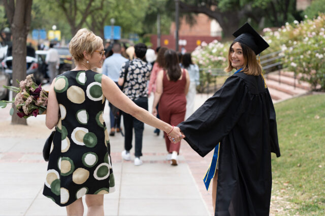 photo - University of California Los Angeles Graduate Wearing Cap and Gown Looking over her Shoulder at the Camera, Holding Hands with her Mother on Graduation Day