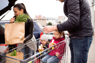 photo - Parents Loading Groceries from Shopping Cart to the Back of the Car and Two Little Daughters Sitting in Shopping Cart Eating Bananas
