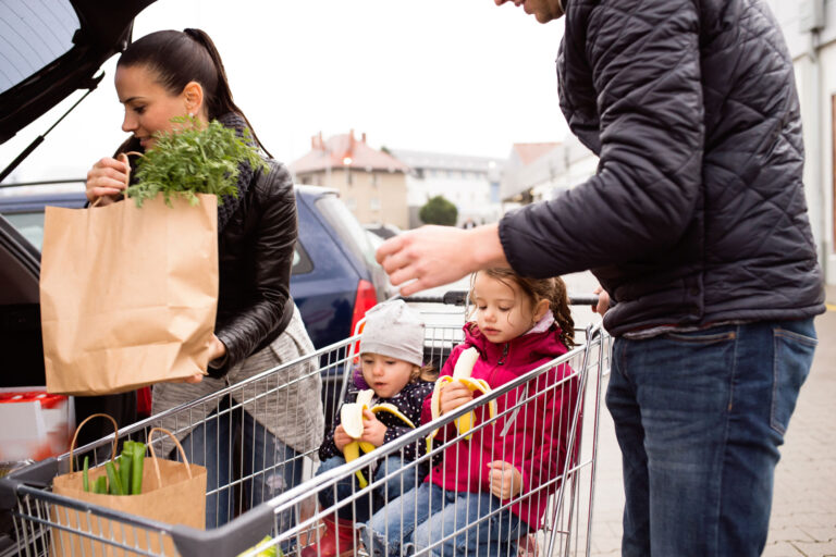 photo - Parents Loading Groceries from Shopping Cart to the Back of the Car and Two Little Daughters Sitting in Shopping Cart Eating Bananas
