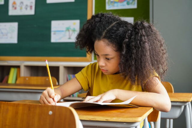 photo - Elementary School Student Sitting at Desk in Classroom and Writing with a Pencil