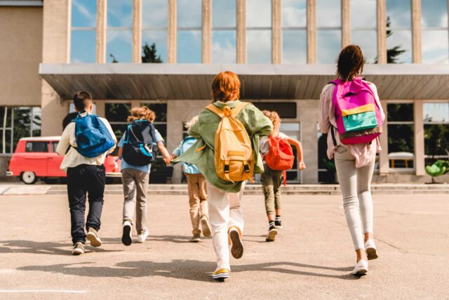 photo - Elementary School Students Walking Towards School and Wearing Backpacks