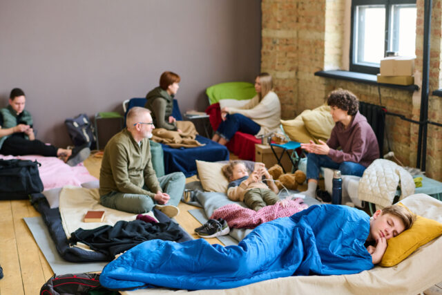 photo - Teenage Boy Sleeping on Cot with Others in Background at Homeless Shelter