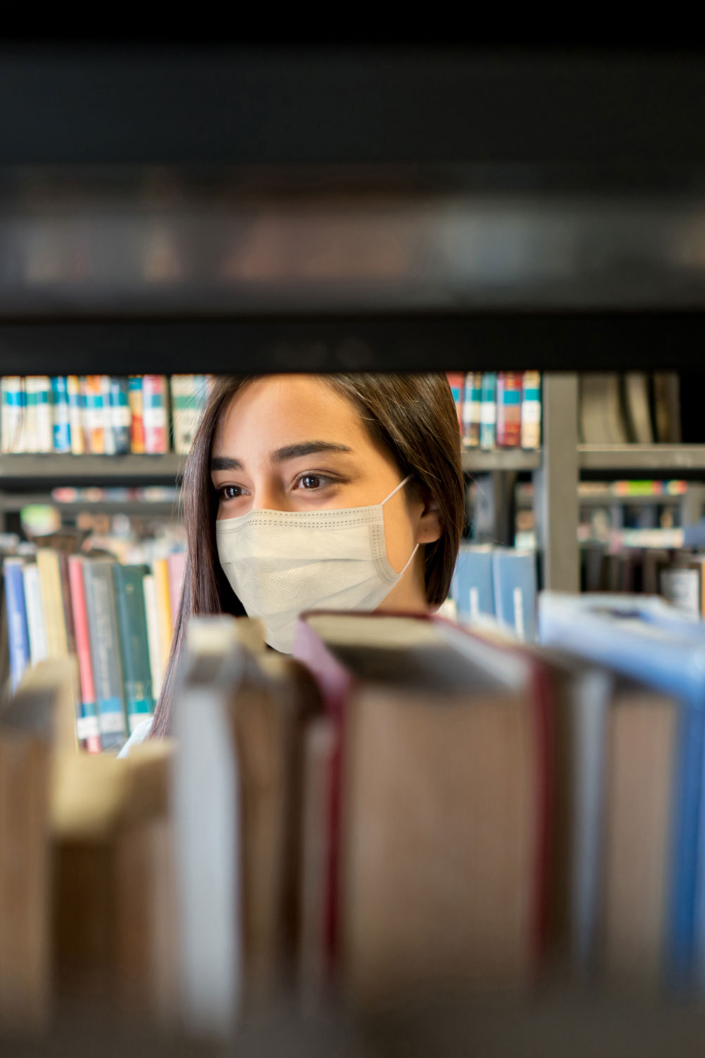 photo - Student Wearing a Facemask at the Library While Looking for a Book