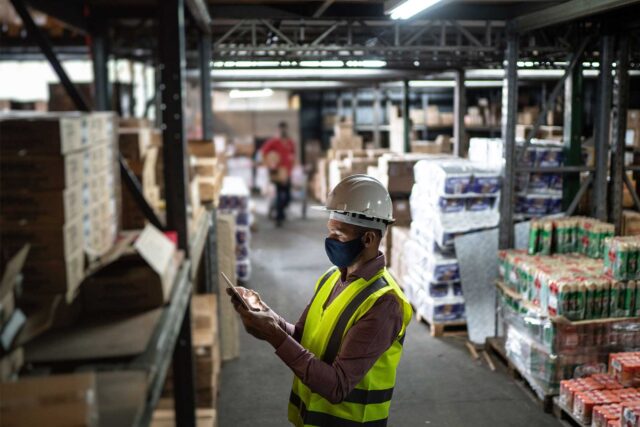 photo - Warehouse Worker Wearing Face Mask and Protective Workwear Checking Products Using Tablet